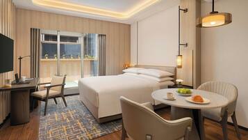 A hotel room with a large bed, a desk with a laptop, a dining table with a plate of food, and a view of the city through the window.