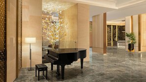 Un piano de cola en un vestíbulo amplio con suelo de mármol, una lámpara moderna y una escultura de árbol decorativa.