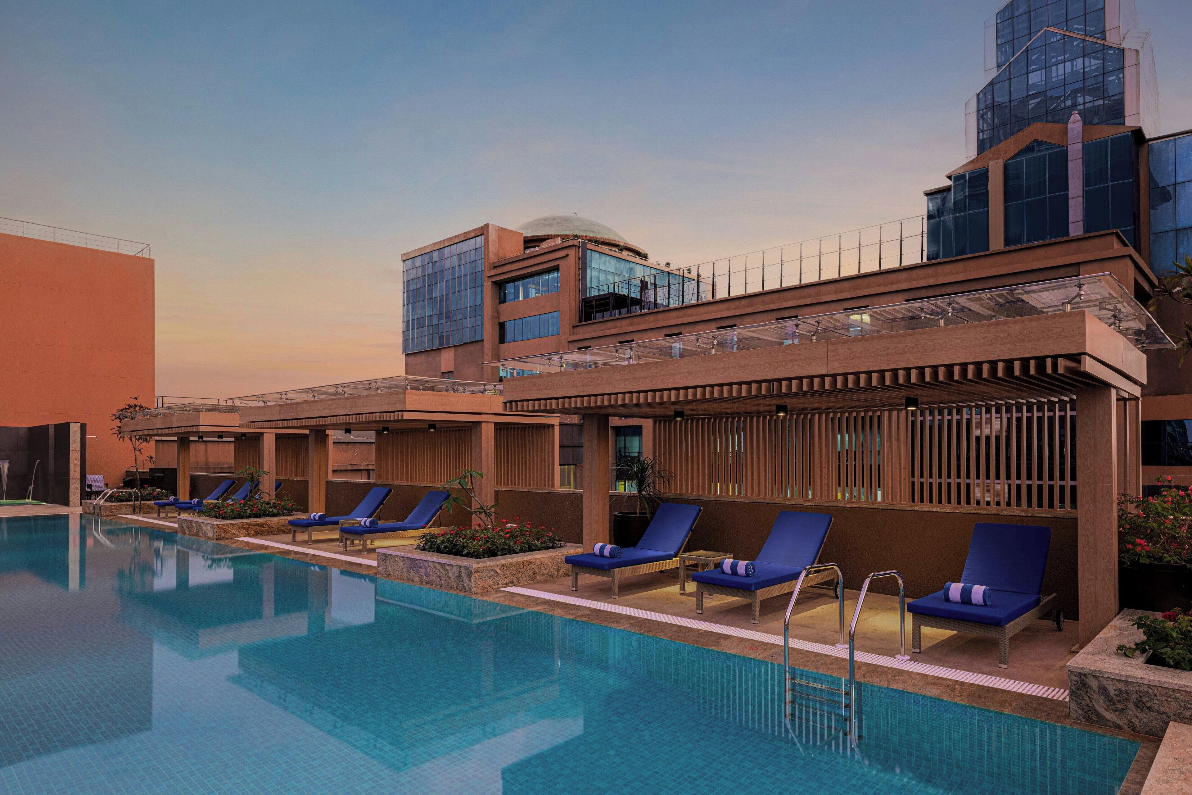 A modern hotel with a swimming pool, lounge chairs, and a multi-story building with large windows.