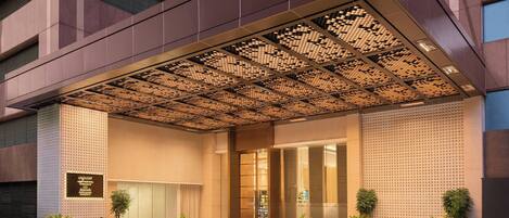 A modern hotel entrance with a geometric patterned canopy, a checkered floor, and potted plants.