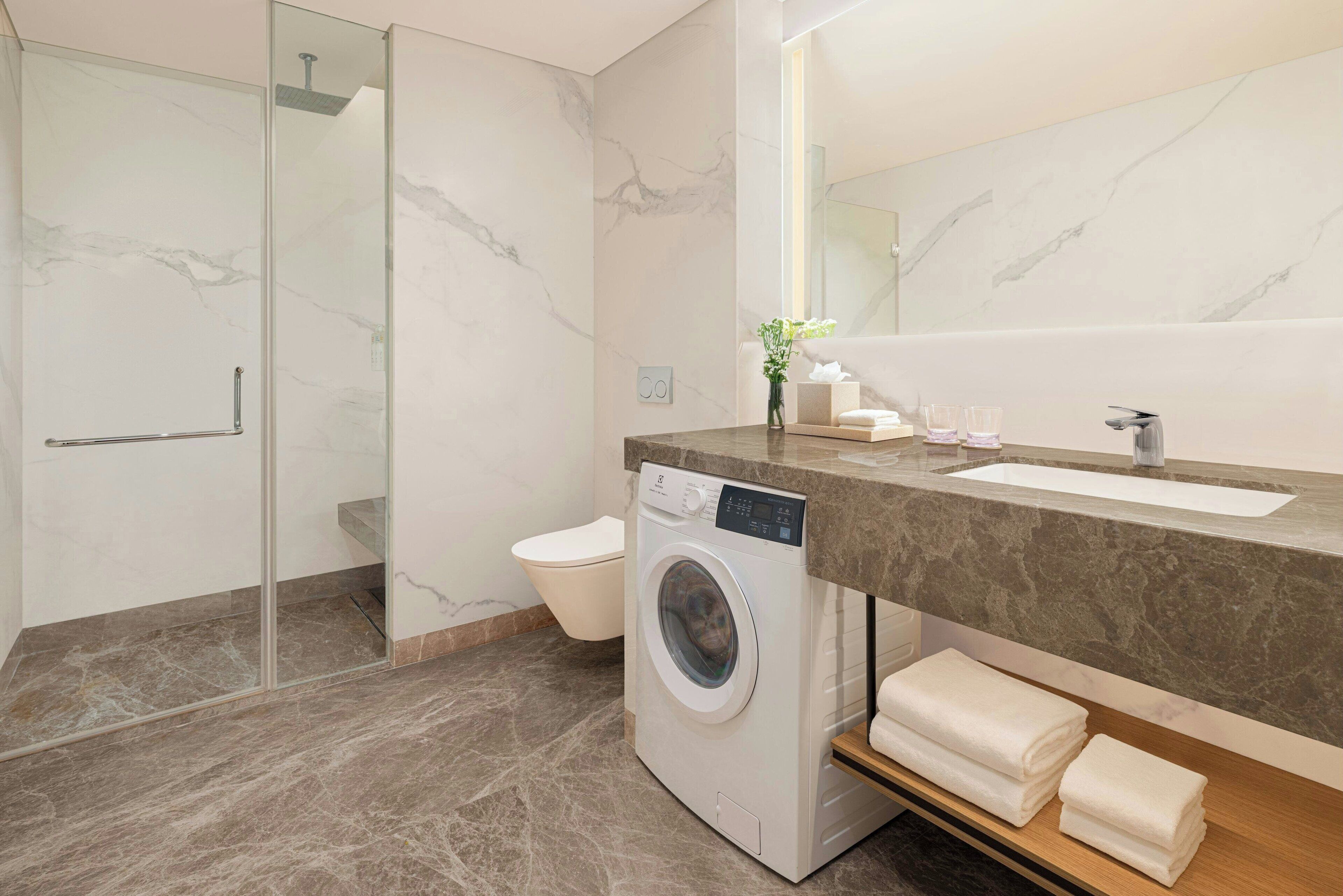 Een moderne badkamer met een marmeren vloer, een glazen douchecabine, een wasmachine, een wastafel met een marmeren aanrechtblad en een spiegel.