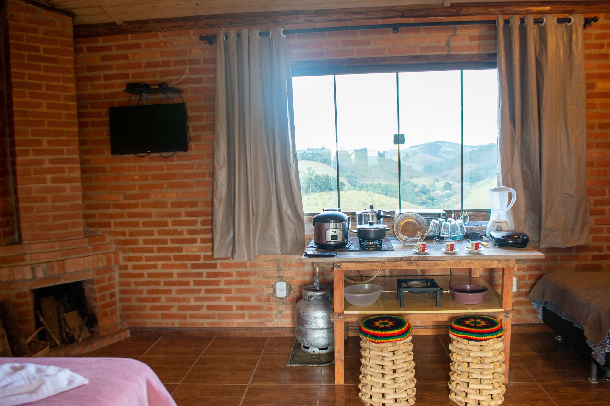 Una habitación rústica con chimenea de ladrillo, un ventanal con vistas a las colinas, una mesa de madera con electrodomésticos de cocina y una cama con una colcha rosa.