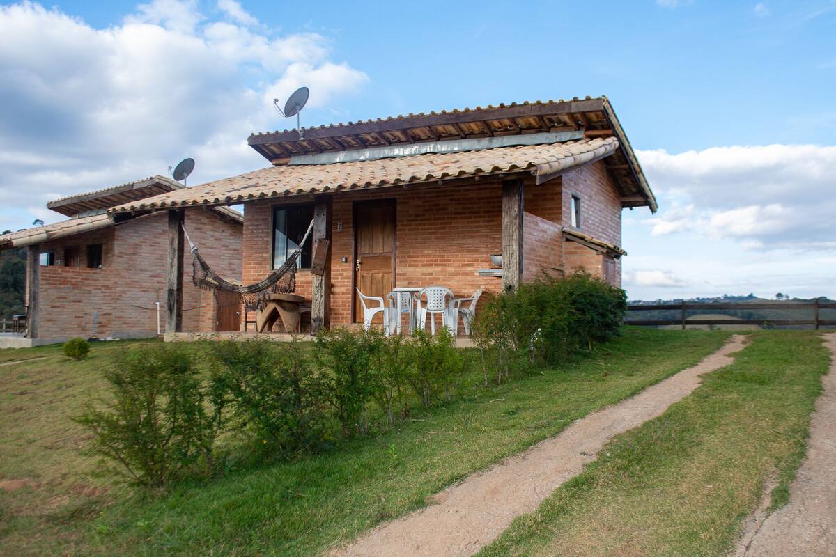 A rustic brick house with a tiled roof, a wooden door, and a small patio with white plastic chairs.