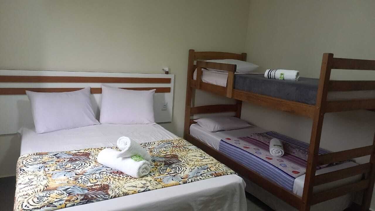 Uma cama de solteiro com um edredom estampado e duas almofadas, uma cama beliche de madeira com um cobertor dobrado e uma pequena mesa com uma toalha.