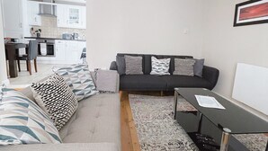 Una sala moderna con un sofá, una mesa de centro y una alfombra. Se ve una cocina al fondo con armarios blancos y techo de madera.