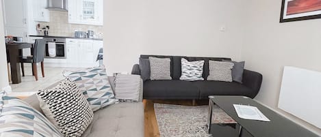 Ein modernes Wohnzimmer mit einer Couch, einem Couchtisch und einem Teppich. Im Hintergrund ist eine Küche mit weißen Schränken und einer Holzbalkendecke zu sehen.