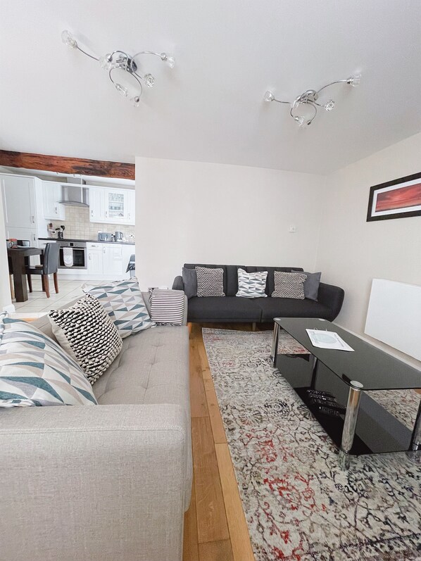A modern living room with a sofa, a coffee table, and a rug. There is a kitchen in the background with white cabinets and a wooden ceiling. - ST ANNAS COTTAGE - wonderful family seaside cottage next to Porthmeor beach (St Ives)