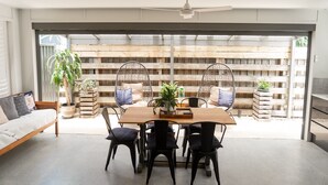 A modern interior with a dining area featuring a wooden table and black chairs, a sofa with cushions, and a view through a glass door to an outdoor space with wooden fencing and plants.