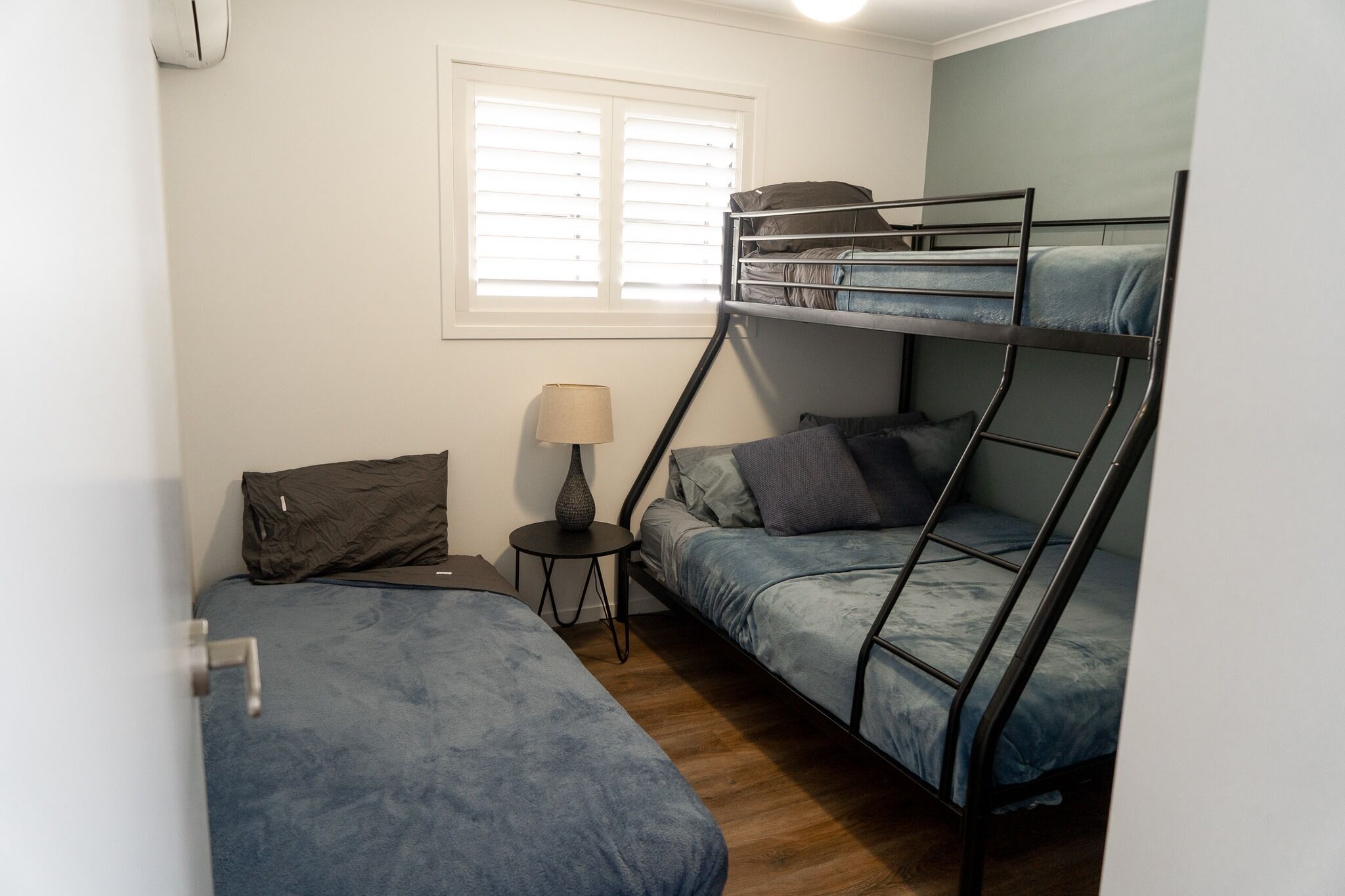 A dormitory room with bunk beds, a small bedside table with a lamp, and a window with blinds.