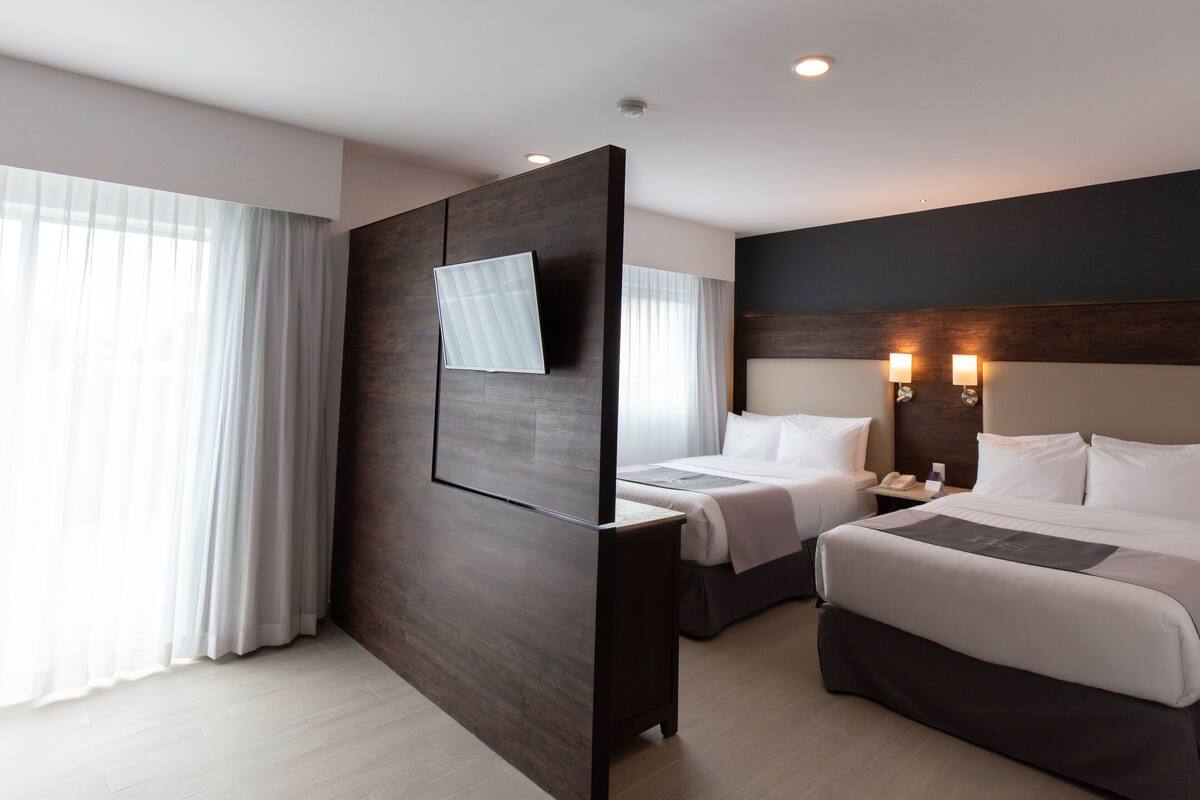 A modern hotel room with a large bed, a smaller bed, a desk, and a TV mounted on a wooden partition.