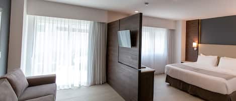 Una habitación de hotel moderna con una cama grande, un sofá, un televisor montado en una partición de madera y ventanas amplias con cortinas translúcidas.