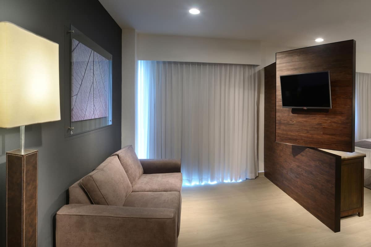 A modern hotel room with a sofa, two beds, a flat-screen TV, and a large window with curtains.