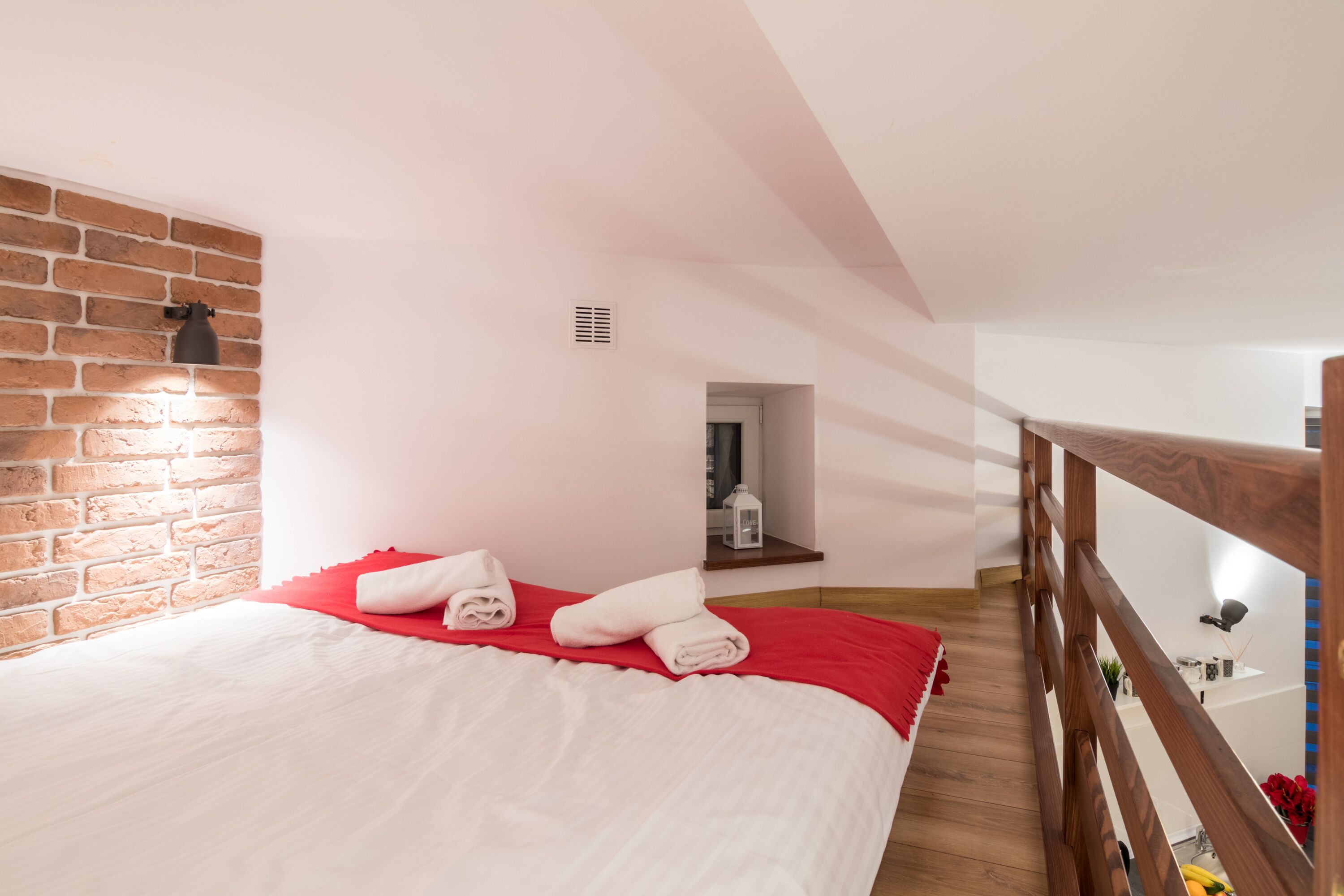 Une chambre avec un mur en briques, un plancher en bois, un lit avec une literie blanche et des touches de rouge, une petite fenêtre et une rambarde en bois.