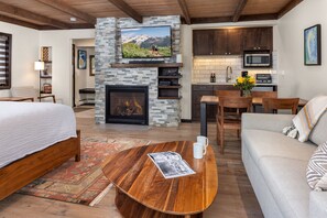 A modern living room with a fireplace, a television mounted on a stone wall, a dining area with wooden furniture, and a kitchen with dark cabinets and a microwave. - Willows Condominiums - C3 (Snowmass Village)