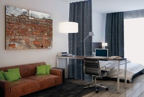 Quarto de hotel moderno com uma obra de arte em tijolo, uma escrivaninha com um computador, uma cama e um sofá com almofadas verdes.