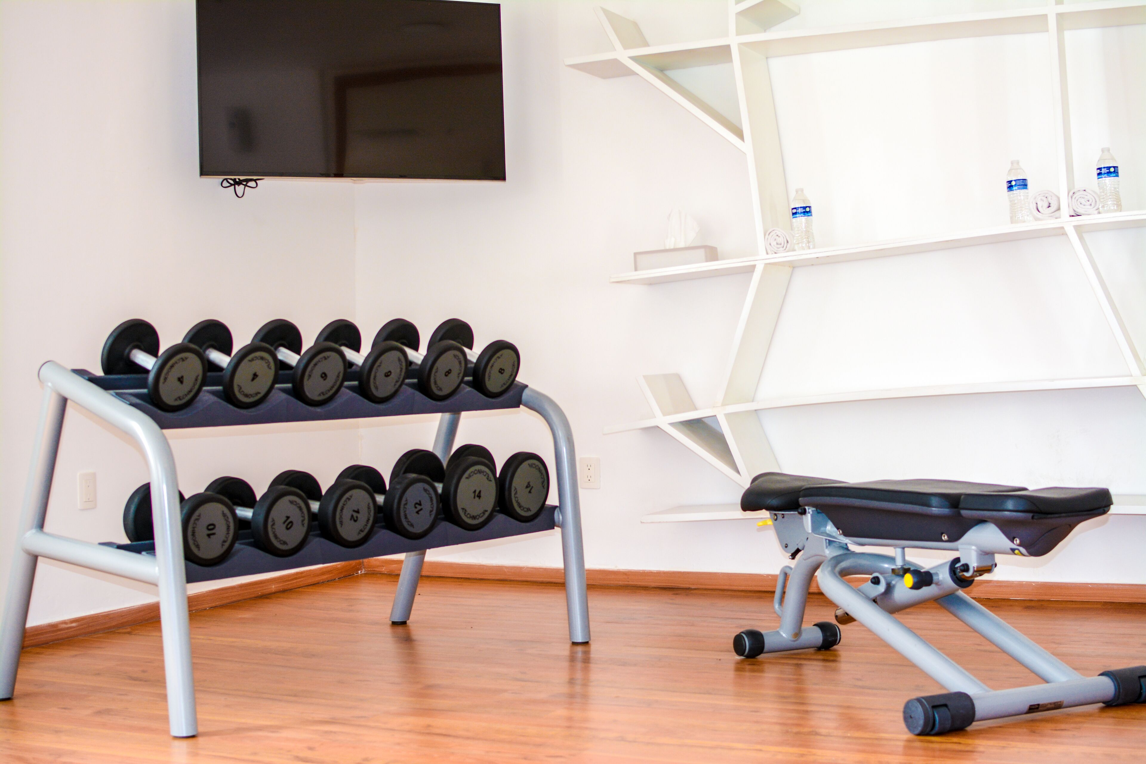 A gym with a dumbbell rack, a bench, and a TV mounted on the wall.