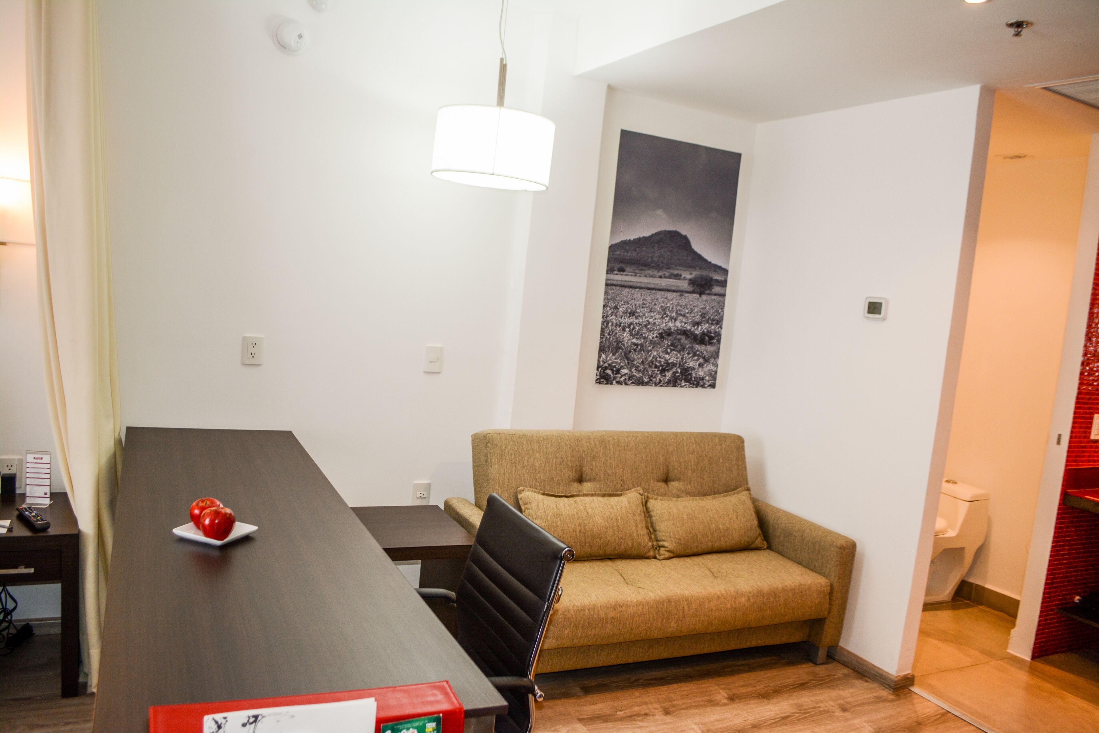 A modern living room with a dark wooden dining table, a beige sofa, and a black chair. There is a white lamp hanging above the table and a black and white photograph on the wall.