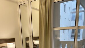 Ein Hotelzimmer mit Bett, Nachttisch mit Lampe, Spiegel, Fenster mit Blick auf Gebäude und Balkon mit Geländer.