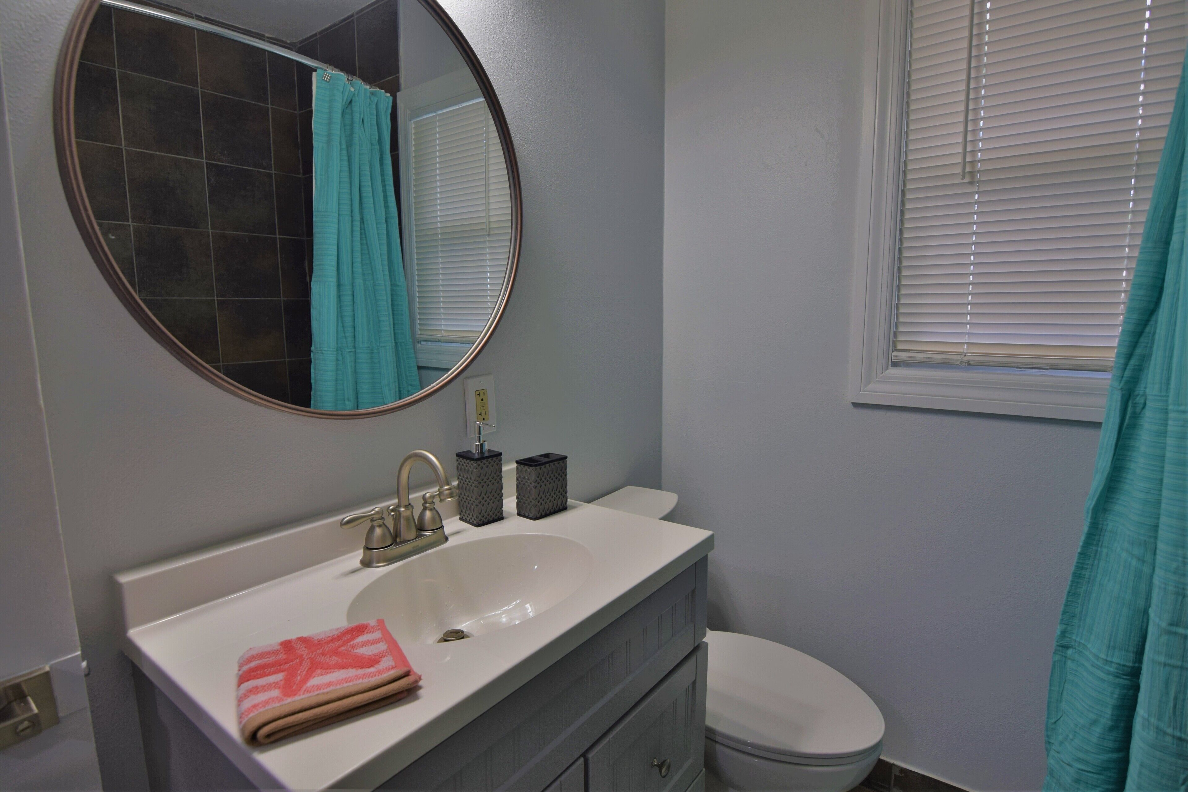 A bathroom with a white sink, a round mirror, a toilet, and a window with blinds.