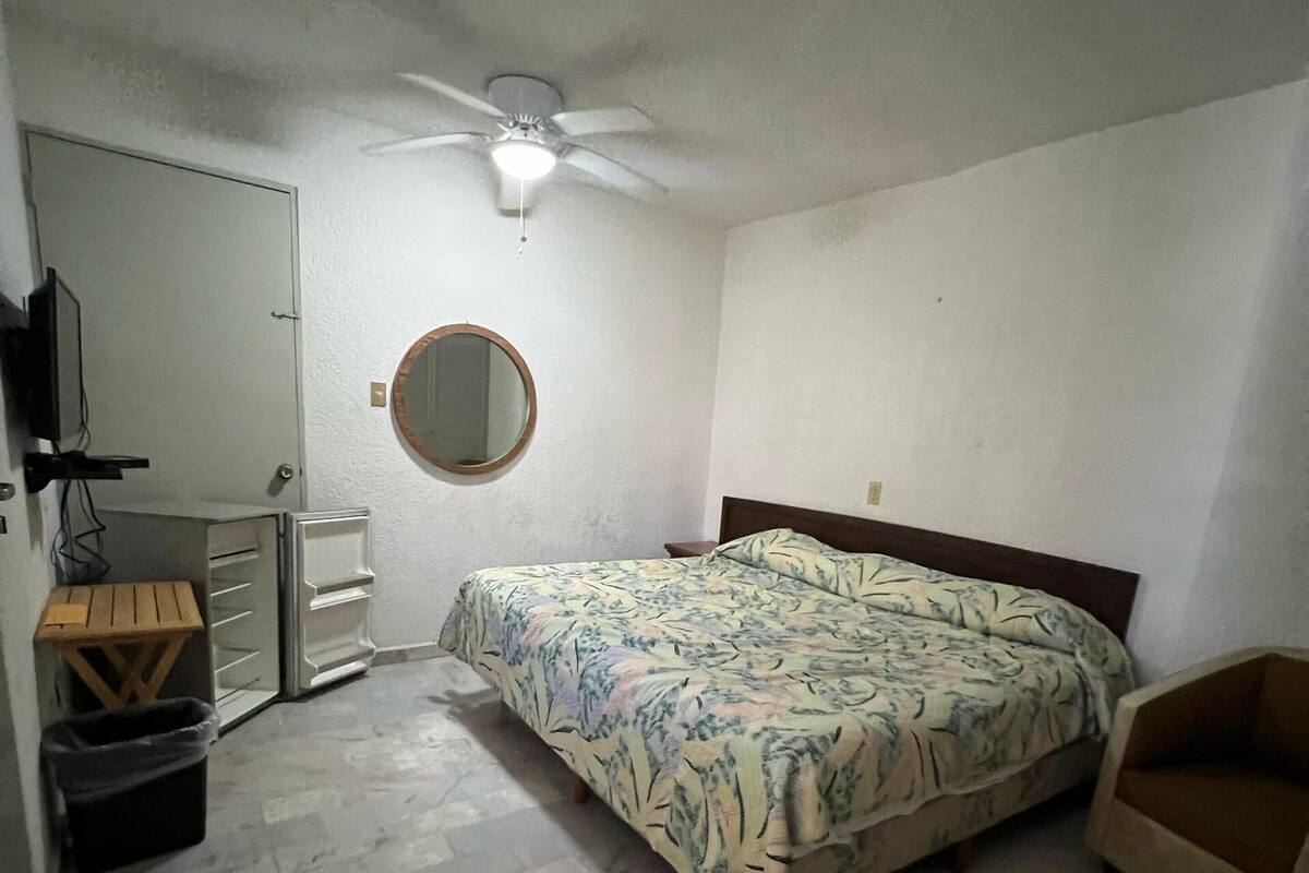 Ein Schlafzimmer mit einem Bett, einem Deckenventilator, einem runden Spiegel, einem Fernseher, einem kleinen Tisch und einem Papierkorb.
