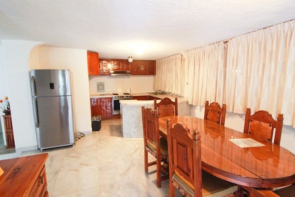 Una cocina con refrigerador de acero inoxidable, armarios de madera, una mesa de comedor con sillas y un ventanal con cortinas.