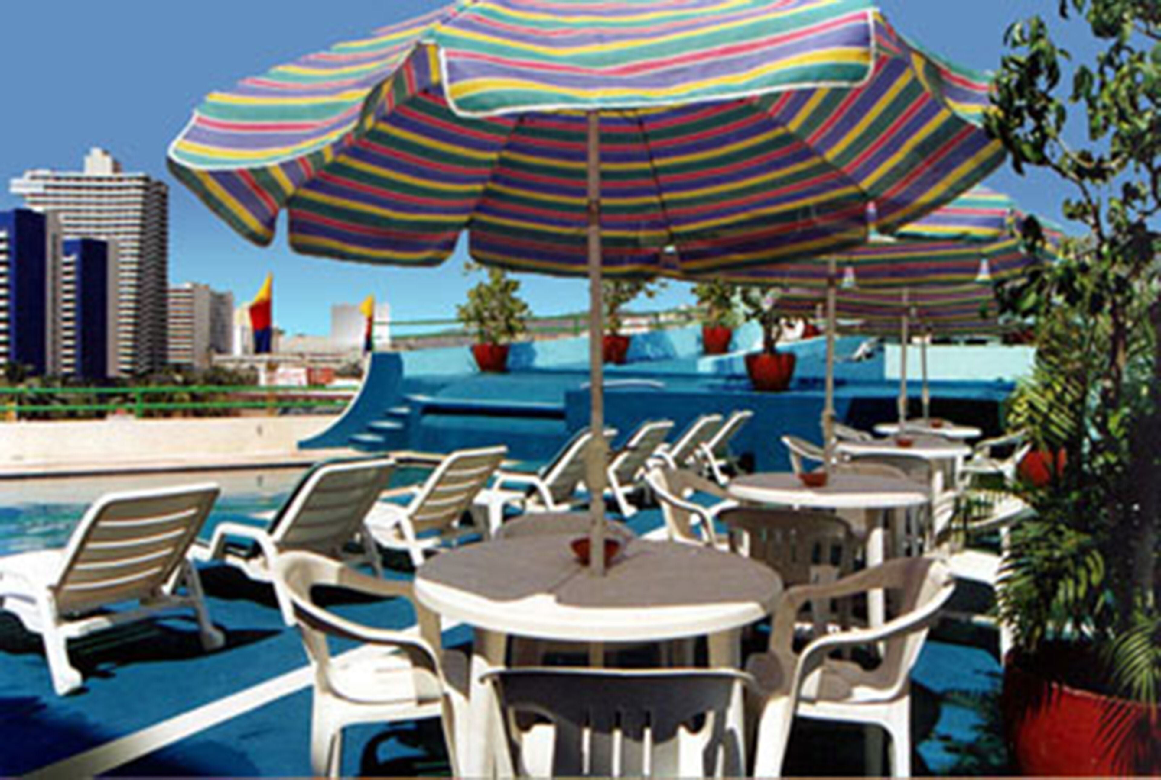 A poolside area with lounge chairs and tables, striped umbrellas, and potted plants.
