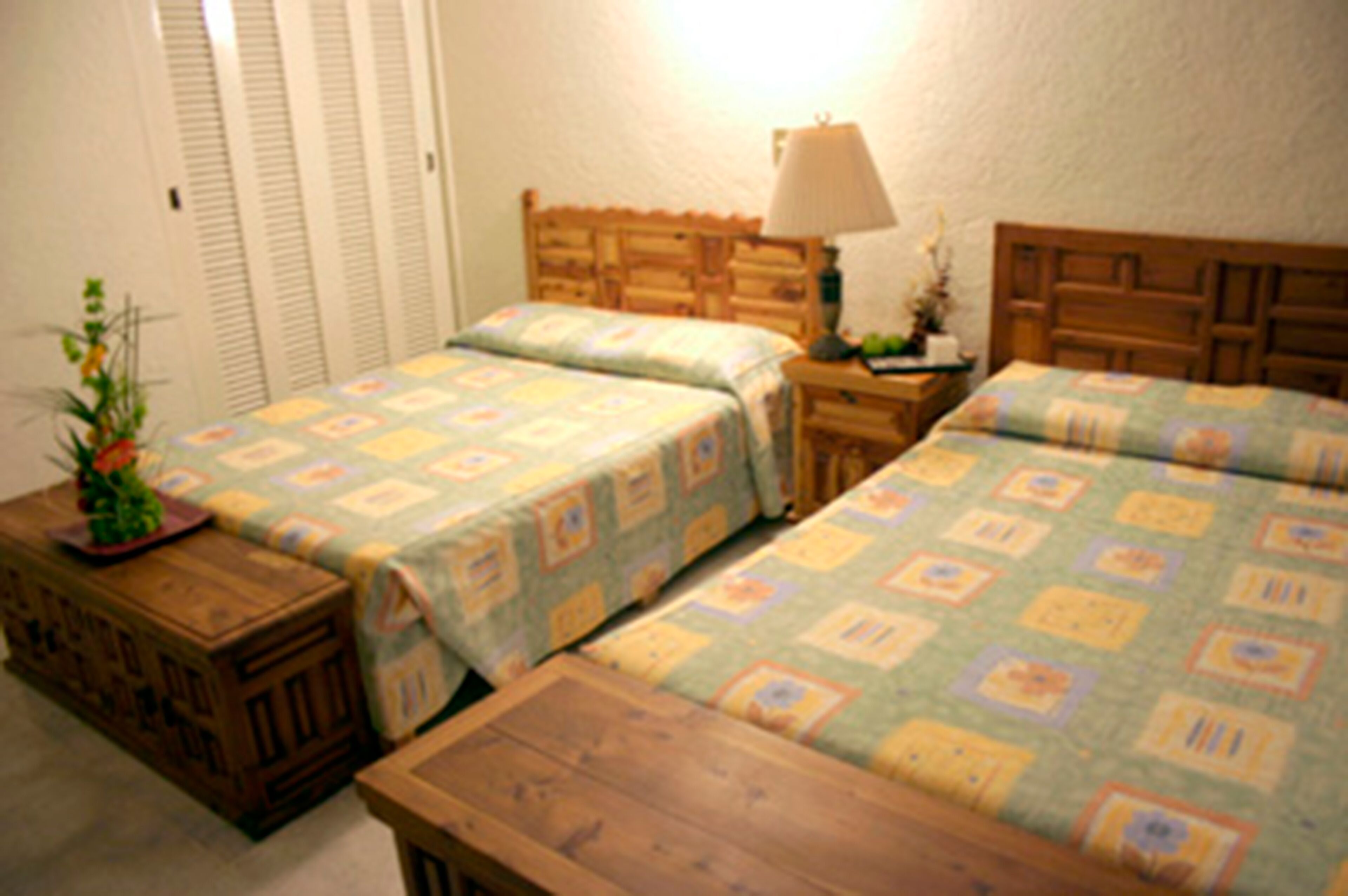 A room with two beds, each with a wooden headboard and a floral-patterned bedspread. There is a nightstand with a lamp between the beds, and a small wooden table with a plant on it.