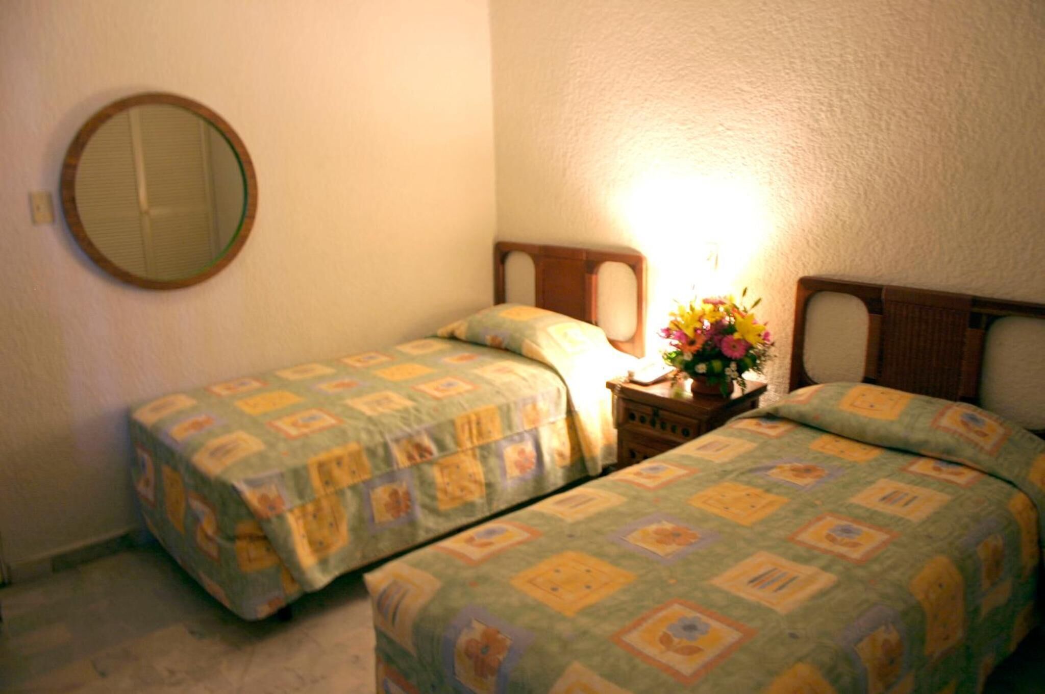 Un dormitorio pequeño y acogedor con dos camas individuales, un espejo redondo, y una mesita de noche con lámpara y flores.