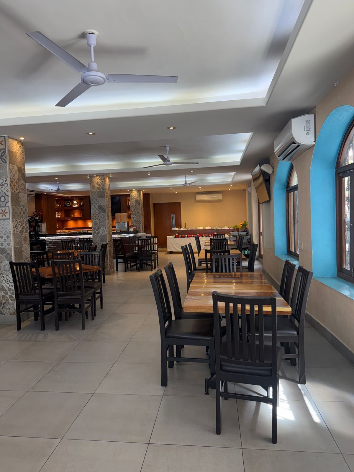 A spacious dining area with multiple tables and chairs, ceiling fans, and a kitchen in the background.
