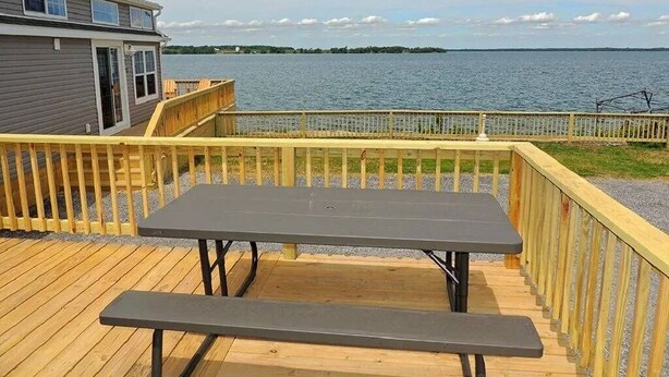 A wooden deck with a picnic table and benches overlooking a body of water. - Cottage #25 - 1 bedroom/ 1 bath (Cape Vincent)