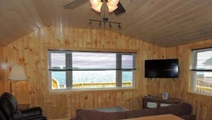 A wooden-paneled room with a large window, a television, a sofa, a recliner, and a wooden coffee table. - Cottage #25 - 1 bedroom/ 1 bath (Cape Vincent)