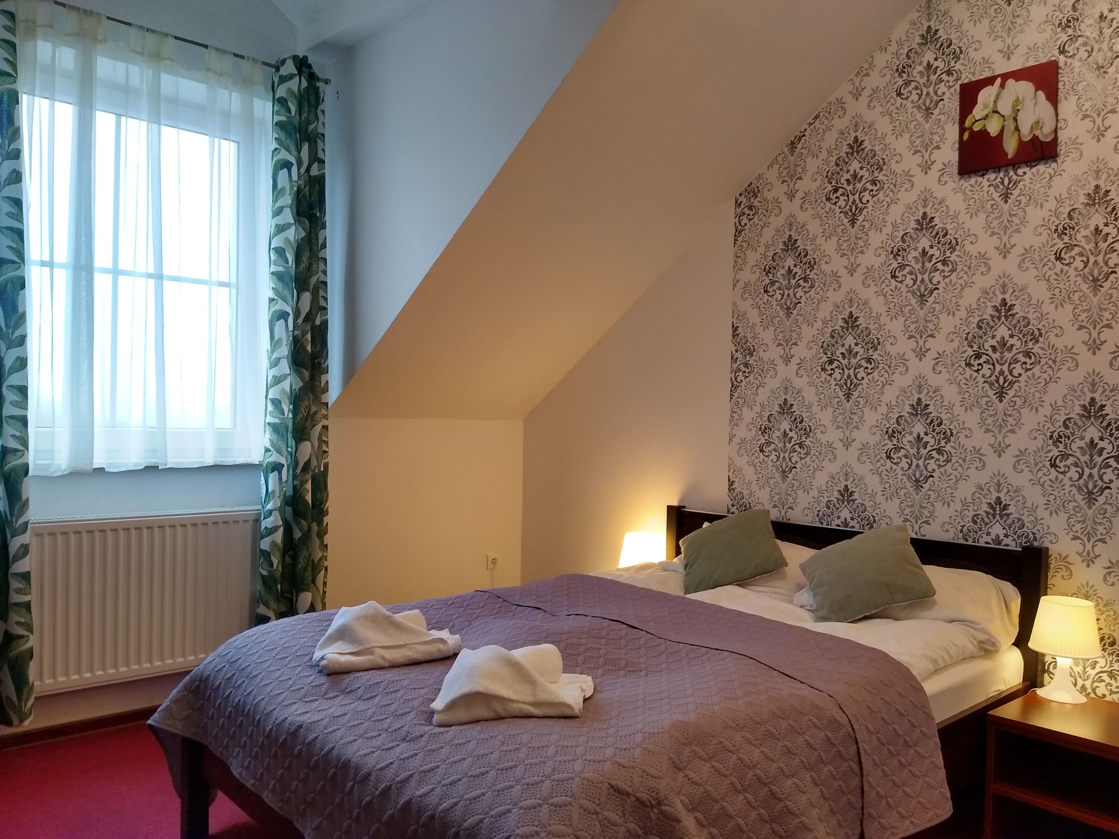 A bedroom with a bed, a nightstand with a lamp, a radiator, and a window with curtains.