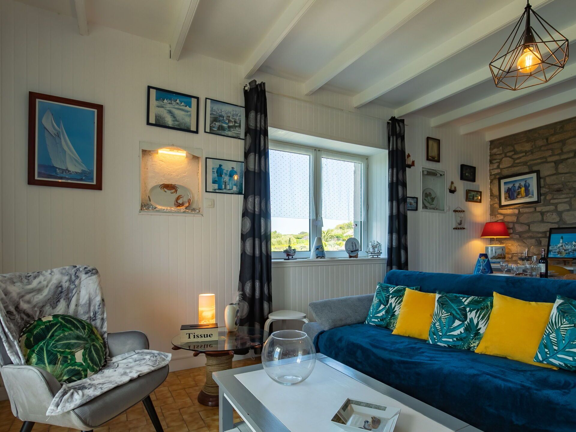 A cozy living room with a blue sofa, a chair with a tropical print cushion, a glass coffee table, and a small side table with a tissue box.
