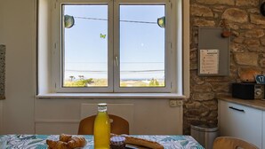 A dining area with a table set for breakfast, including croissants, a bottle of juice, and a baguette. A window with a view of the sea and a stone wall is visible. - Stunning private villa for 4 people with WIFI, TV and pets allowed (Le Vourch)