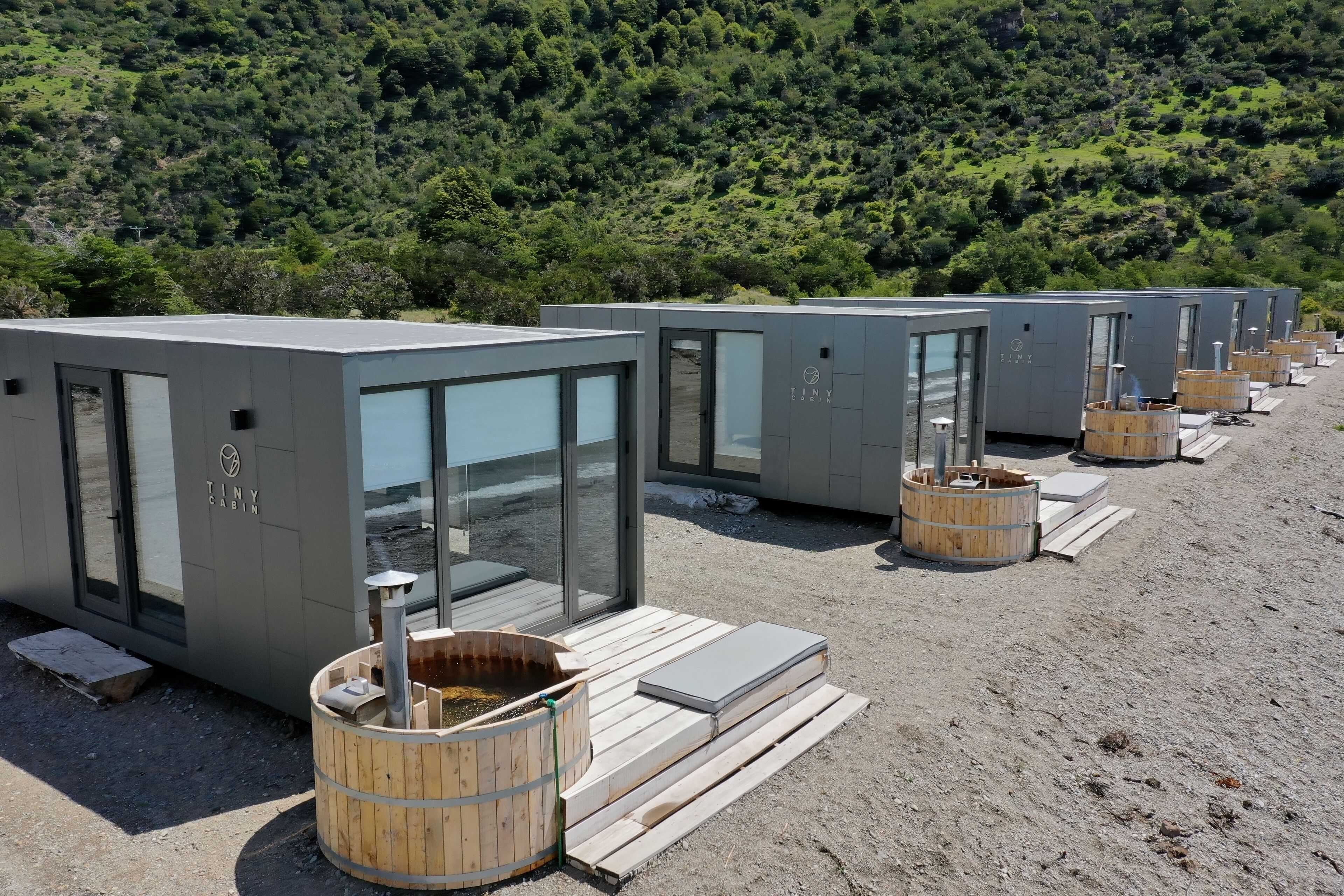 A row of modern cabins with glass doors, wooden steps, and a hot tub in front.