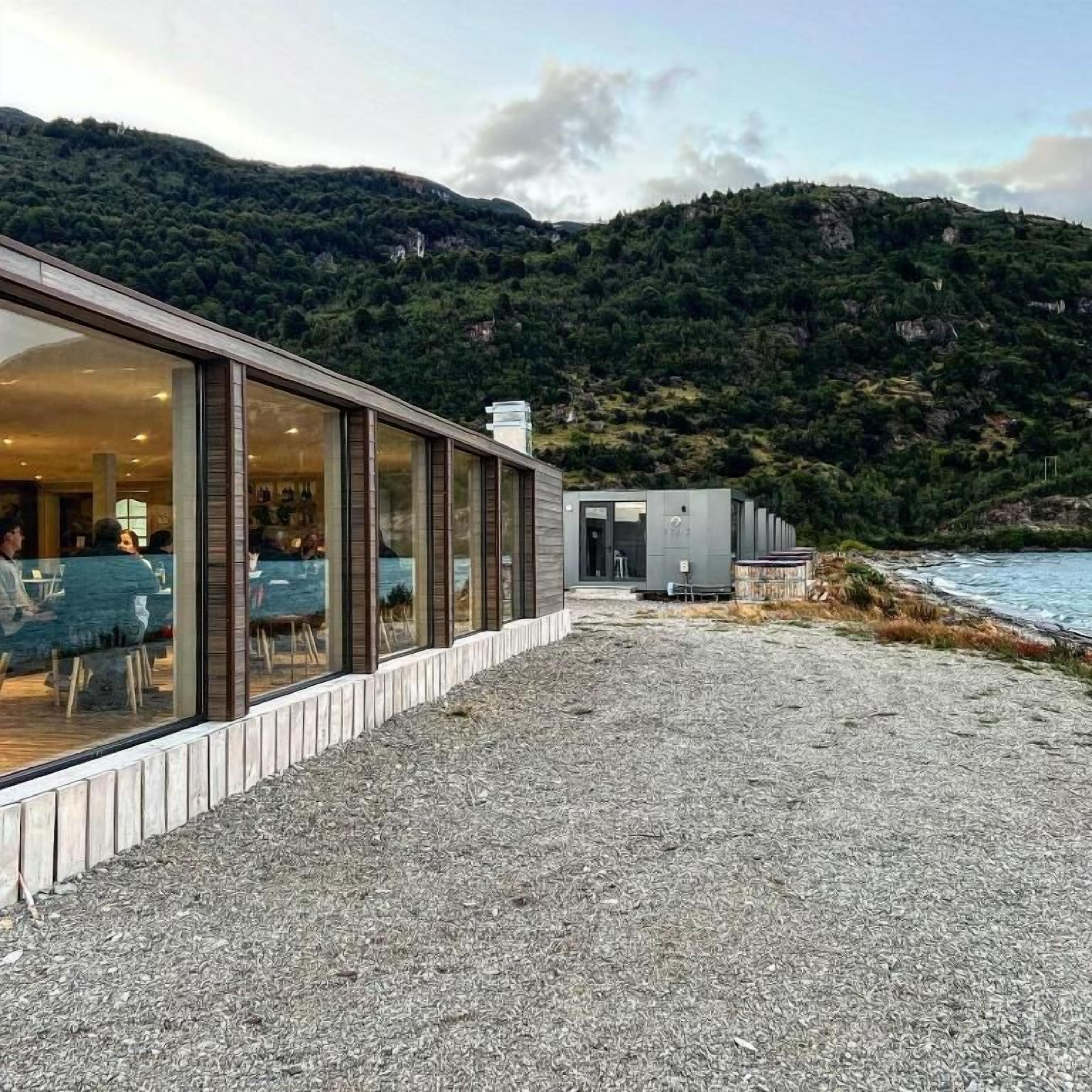 A building with a gravel path leading to a glass-enclosed dining area, overlooking a body of water and a mountainous landscape.