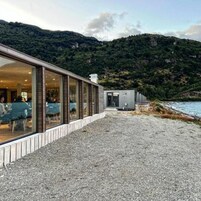 Un edificio con un camino de grava que conduce a un área de comedor con paredes de vidrio, con vista a un cuerpo de agua y un paisaje montañoso.