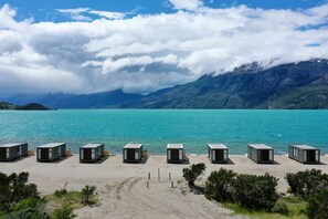 Une rangée de cabanes de plage modernes, de forme rectangulaire, offrant une vue dégagée sur un lac turquoise et sur des montagnes en arrière-plan.