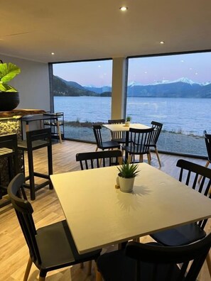 Un coin repas avec une table et des chaises, une plante en pot, et une vue sur un lac avec des montagnes en arrière-plan.