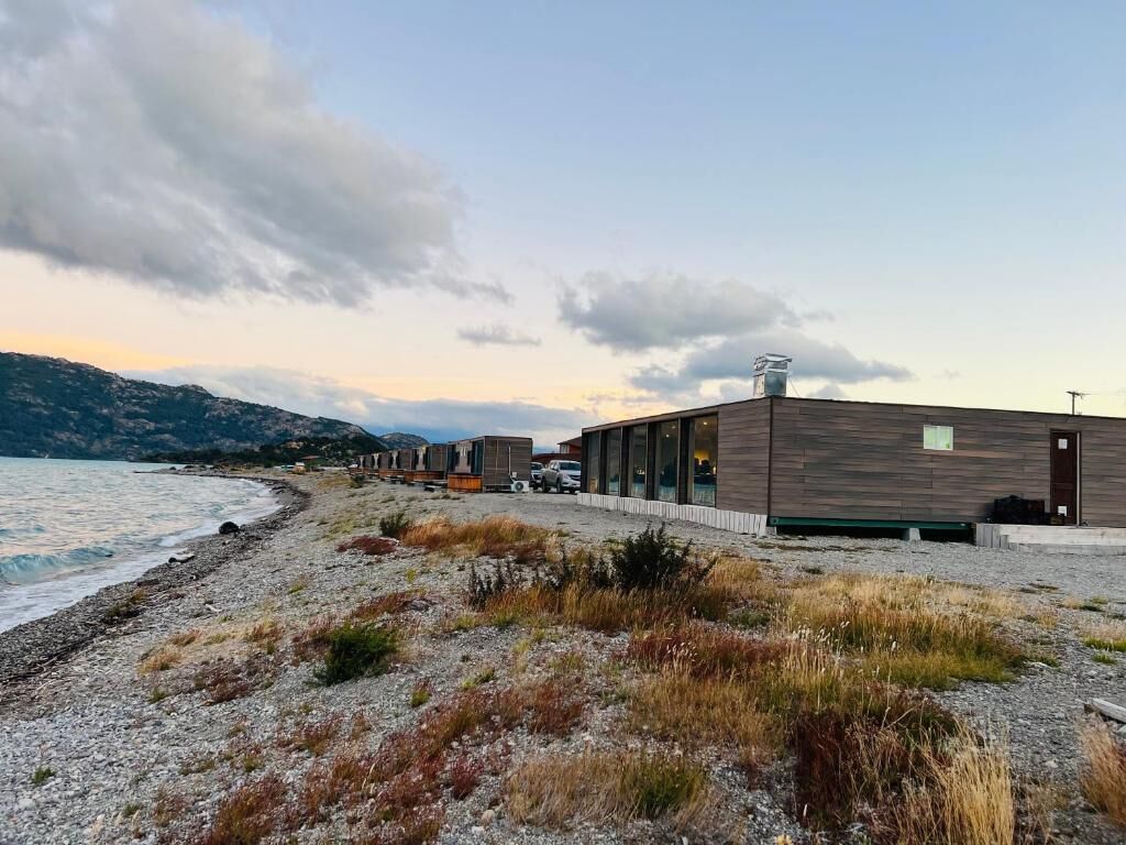 Un edificio moderno con amplios ventanales, ubicado junto a una playa de guijarros y vistas al mar y colinas al lejano.