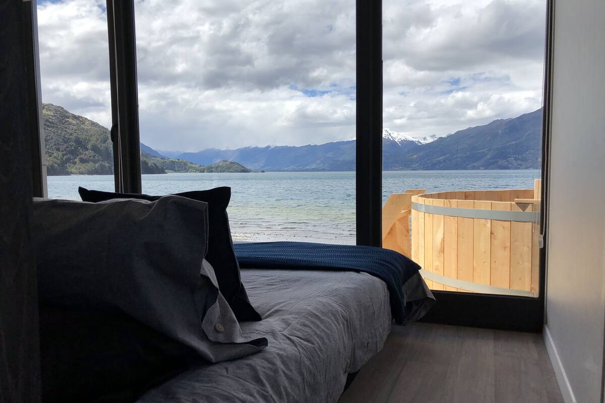 A room with a bed, large windows, and a view of a lake and mountains.