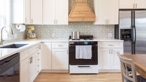 A kitchen with white cabinets, a black stove, a wooden range hood, a stainless steel refrigerator, and a wooden dining table. - Blue Cottage in the heart of Tulsa (Tulsa)