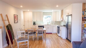 A modern kitchen with white cabinets, a dining area with a wooden table and chairs, a refrigerator, and a bookshelf with books. - Blue Cottage in the heart of Tulsa (Tulsa)