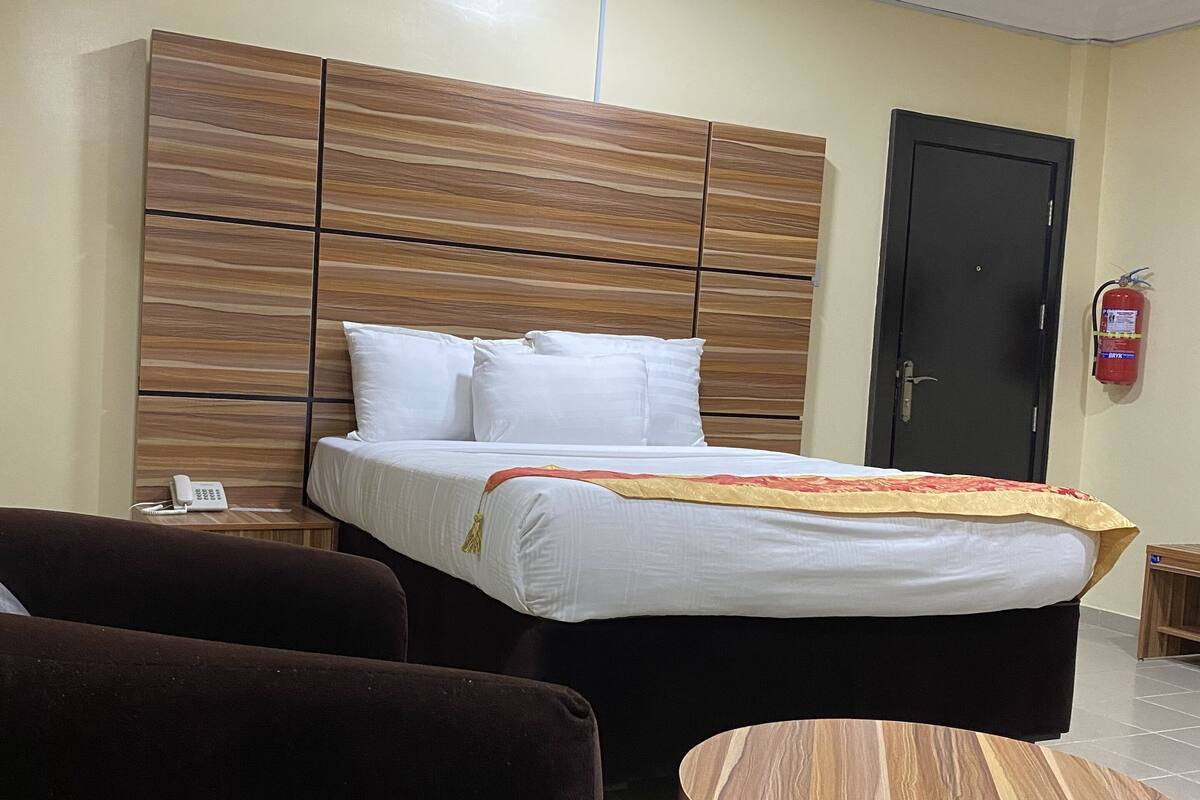 A hotel room with a large bed, a wooden headboard, a bedside table with a phone, a round wooden table, and a dark-colored armchair.