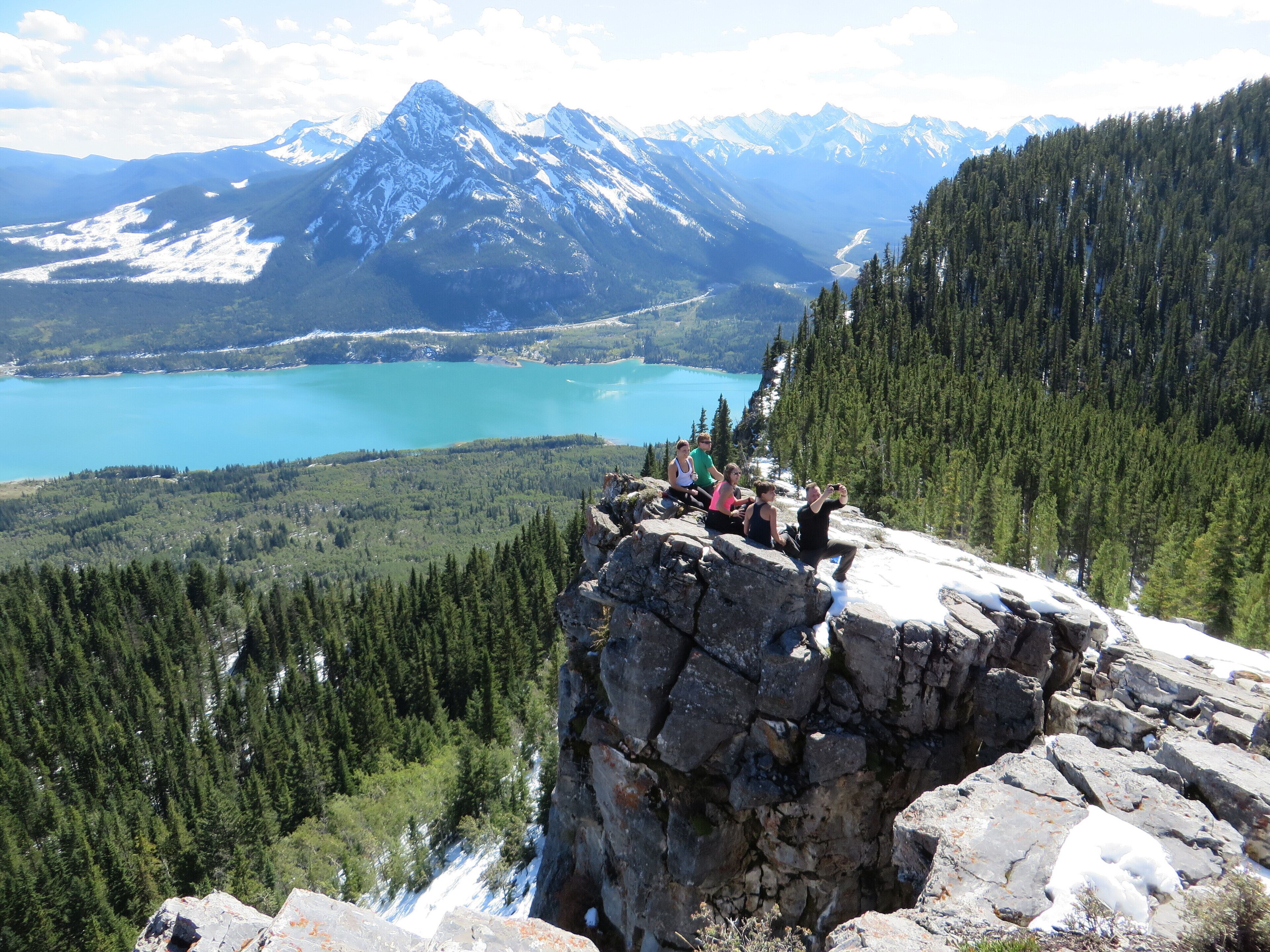 A group of people sitting on a rocky outcrop overlooking a turquoise lake and snow-capped mountains.