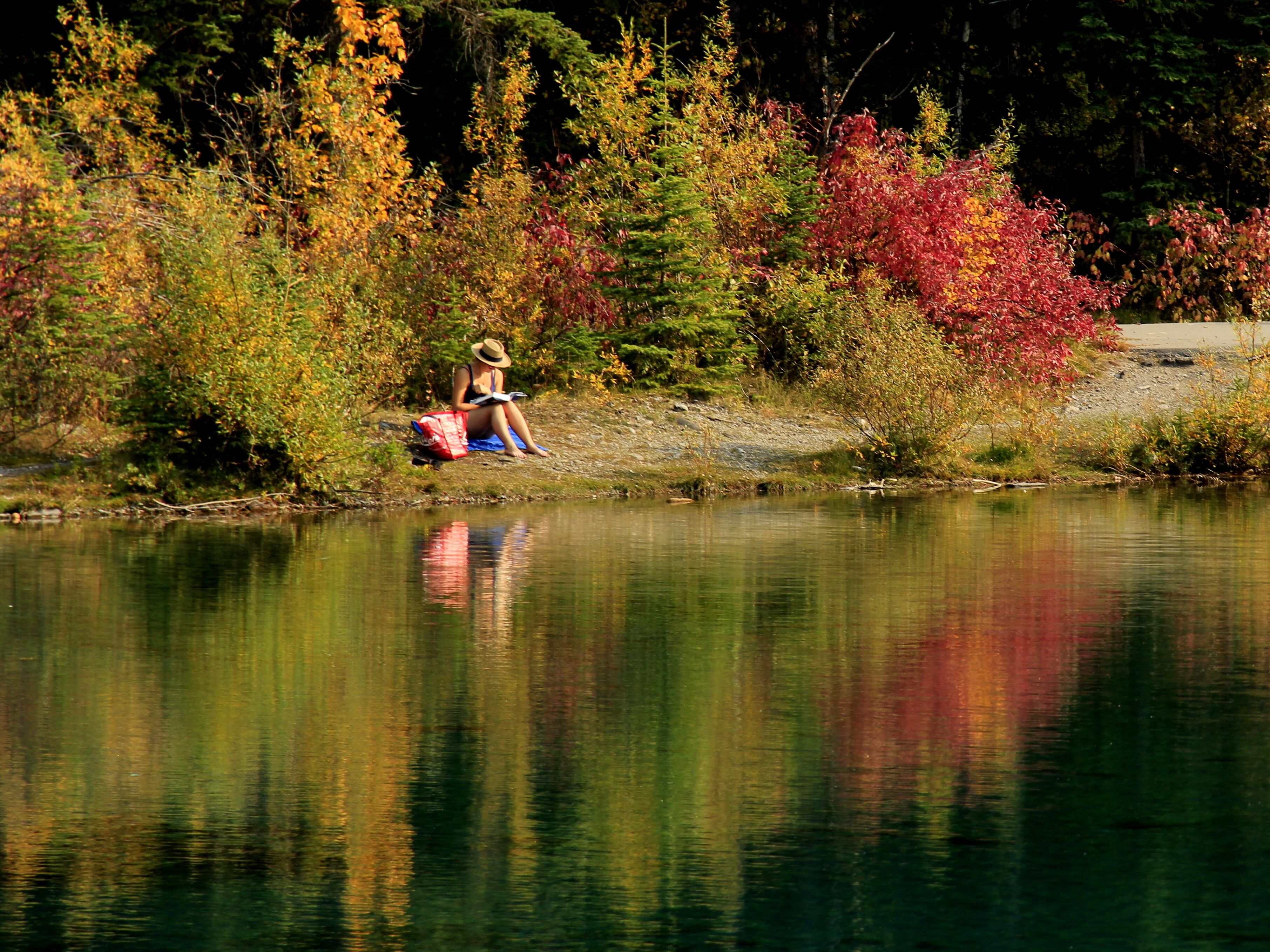 Una persona sentada junto a un lago tranquilo con follaje otoñal al fondo.