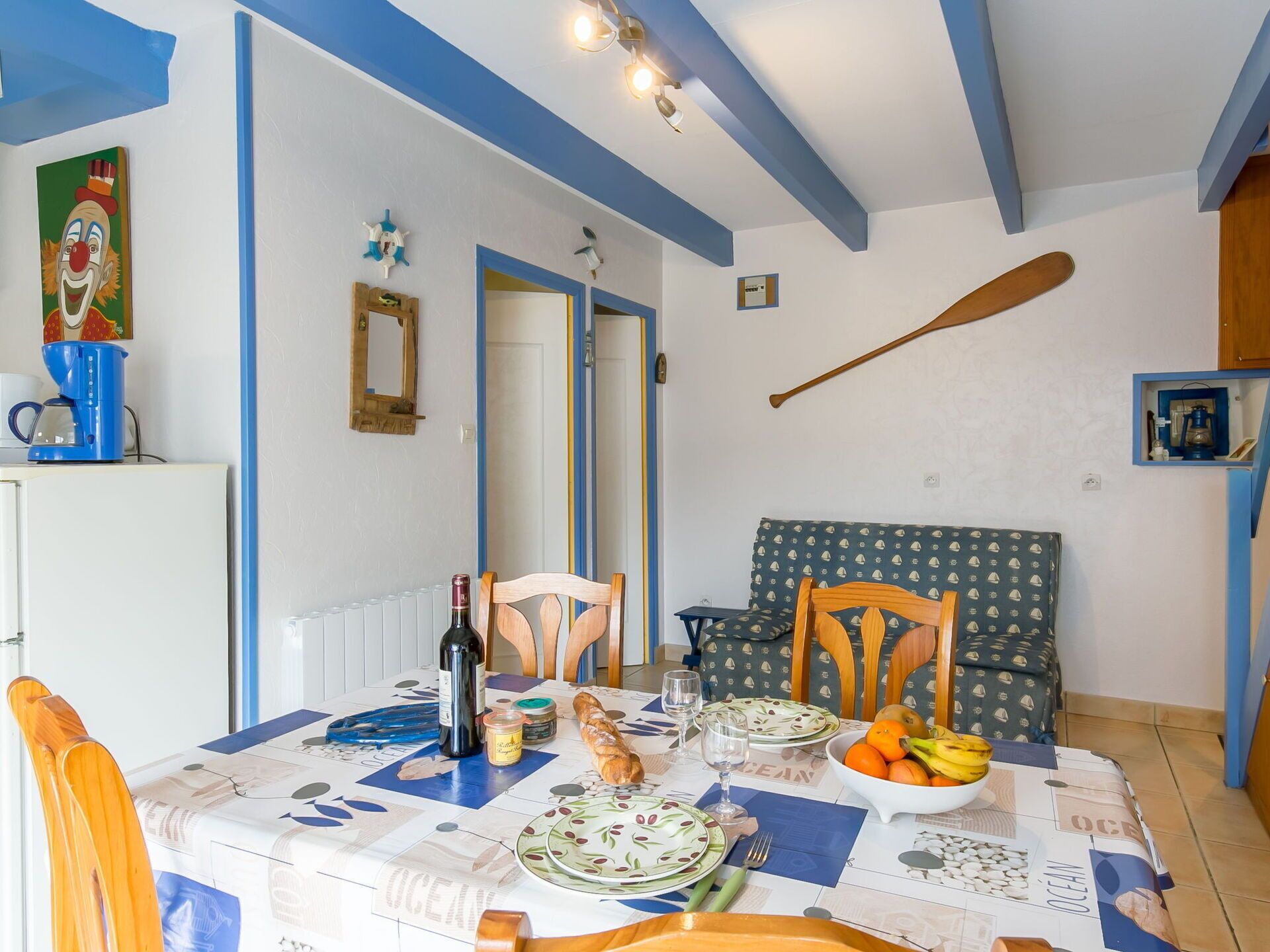 A dining area with a table set for two, featuring a patterned tablecloth, plates, and a bottle of wine. There is a wooden paddle on the wall, a framed picture, and a blue coffee maker on the counter.