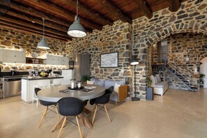 Une cuisine spacieuse avec un mur en pierre, des poutres en bois au plafond, un coin repas avec une table ronde et des chaises, et un escalier menant à un niveau supérieur.