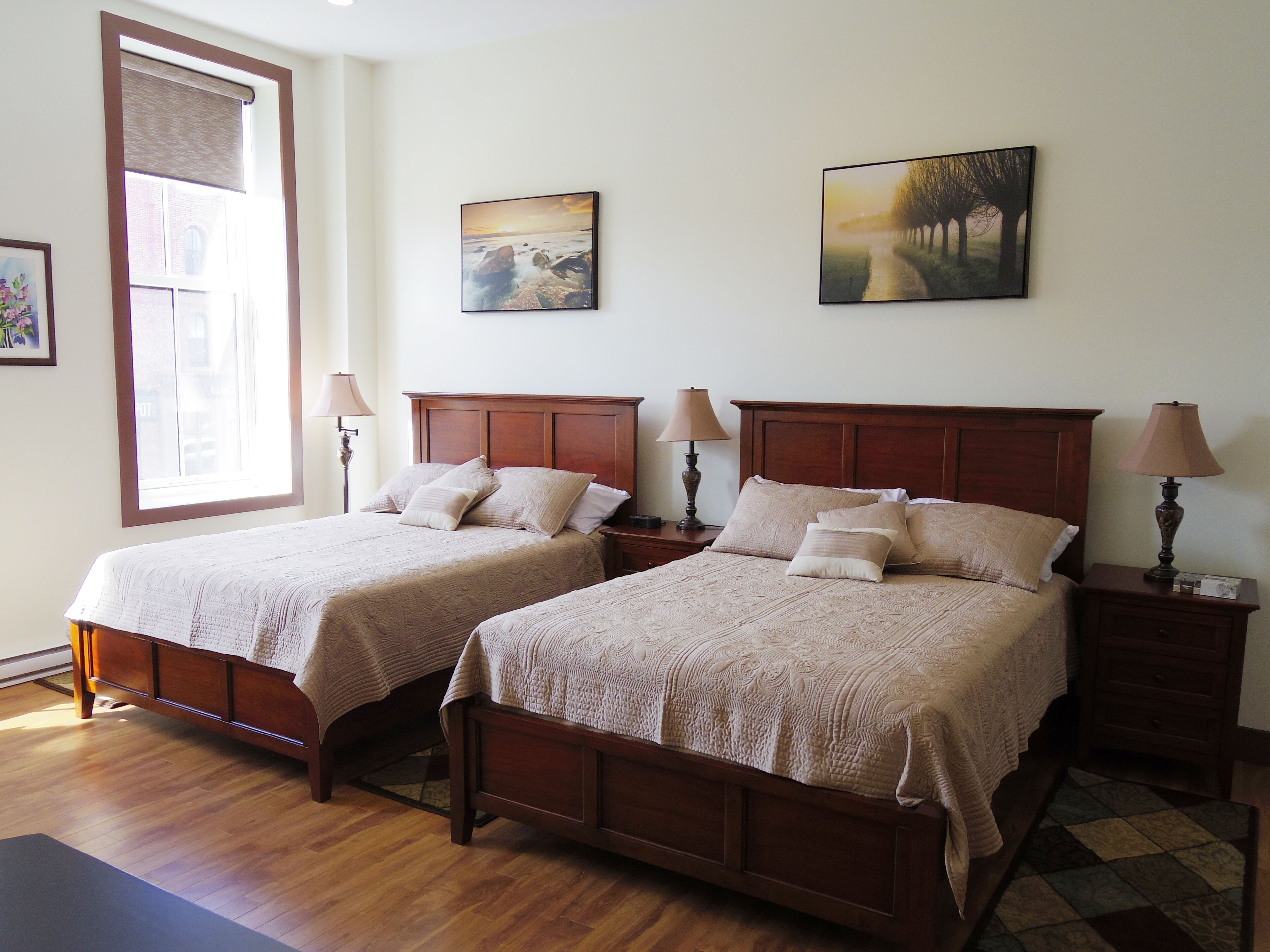 Une chambre d’hôtel avec deux lits en bois, chacun muni d’une table de chevet et d’une lampe. Il y a une fenêtre donnant sur l’extérieur, ainsi que deux tableaux accrochés au mur.