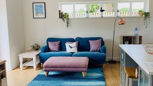 A modern living room with a blue sofa, a wooden coffee table, and a blue rug. There is a window with plants on the sill and a picture on the wall. - Beautiful, stylish New Forest 2 bed cottage, close to the beach! (Lymington)